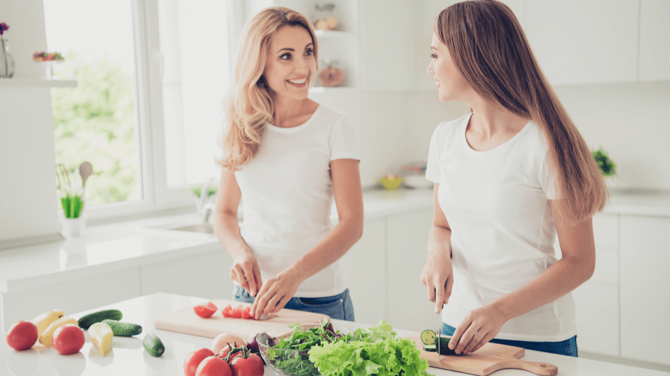 Дві жінки усміхнені на кухні, стоять поруч біля кухонного столу і ріжуть на дошках овочі, попереду розташовані різні непорізані овочі.